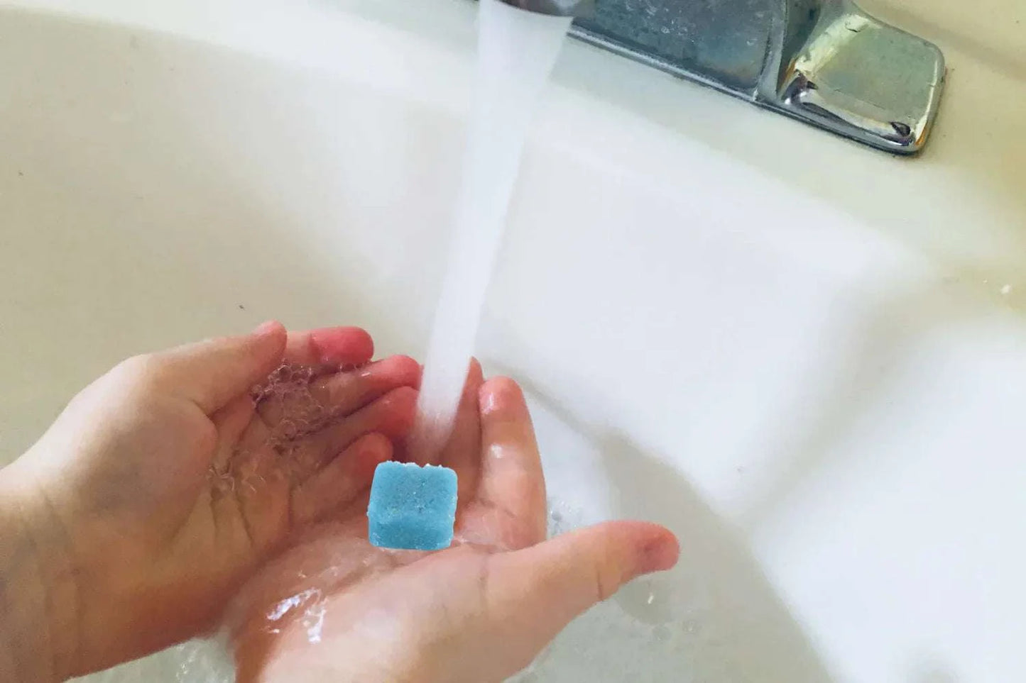 Hands holding a blue sugar scrub cube under running water.