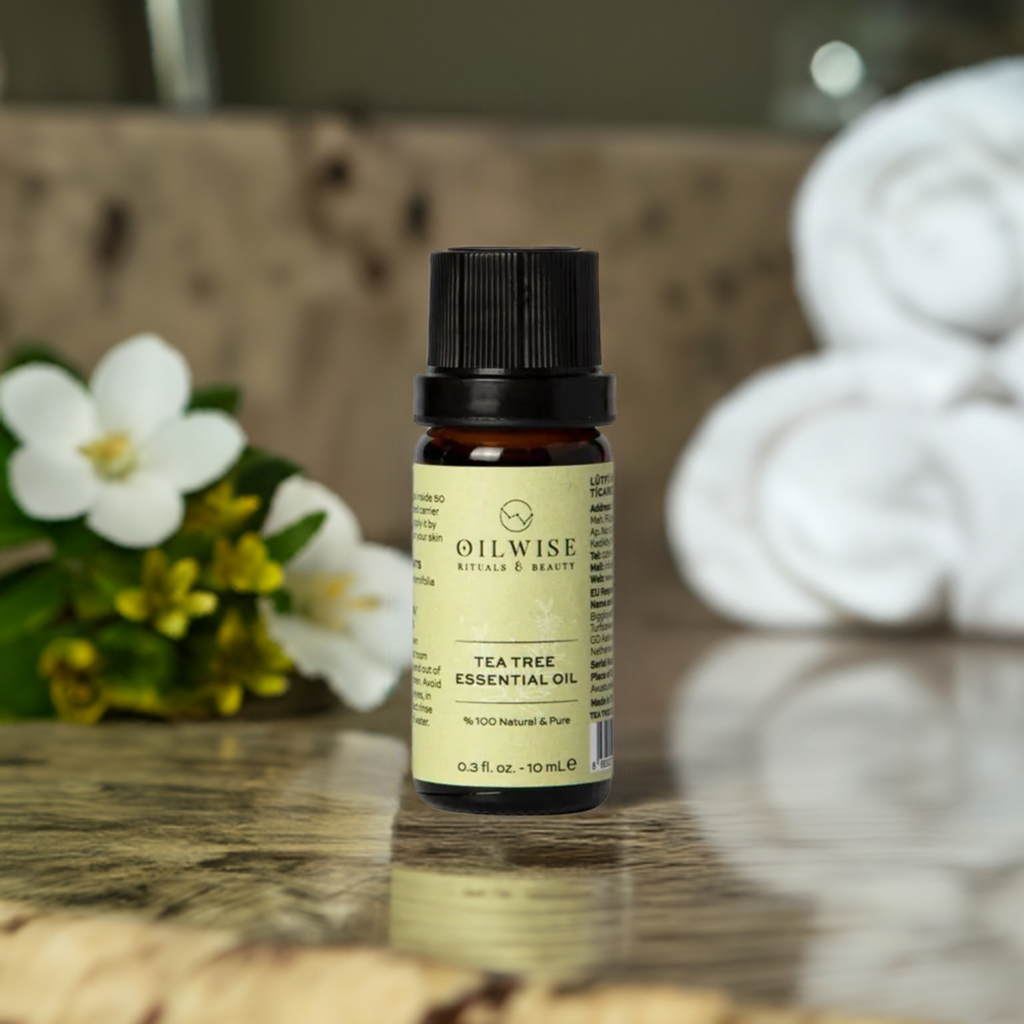 Oilwise Tea Tree Essential Oil bottle on a reflective surface with white flowers and towels in the background.